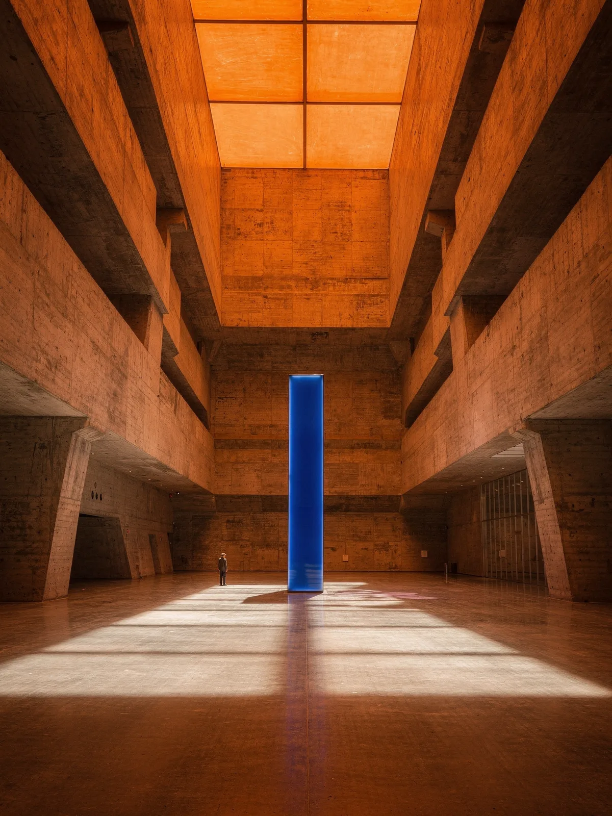 A cavernous concrete atrium bathed in warm orange light, a tall cobalt-blue light column standing at its center beside a lone figure.